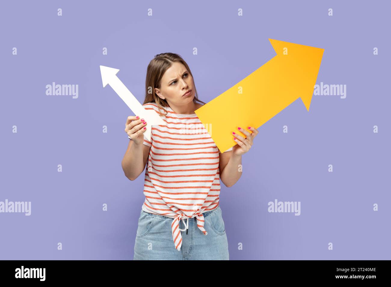 Portrait of confused pensive woman wearing striped T-shirt holding two arrows indicating to different sizes, looking at indicators with puzzlement. Indoor studio shot isolated on purple background. Stock Photo