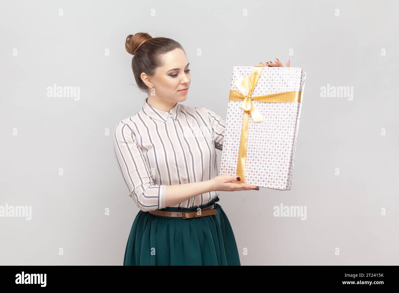 Portrait of young adult beautiful woman wearing striped shirt and green skirt standing showing wrapped present box, congratulating with holiday. Indoor studio shot isolated on gray background. Stock Photo