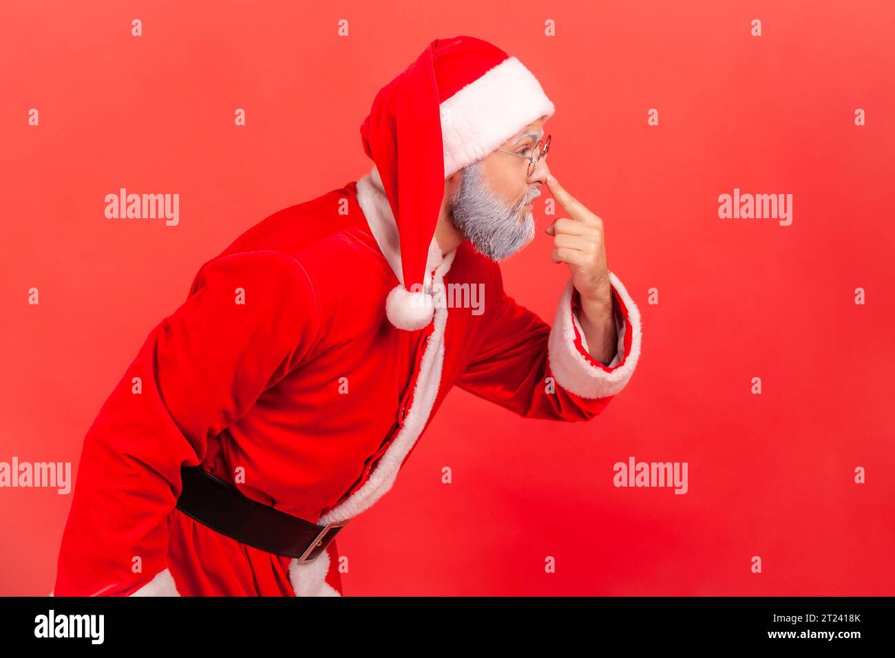 Side view portrait of elderly man with gray beard wearing santa claus costume touching nose with finger, reproaching liar in deception. Indoor studio shot isolated on red background. Stock Photo