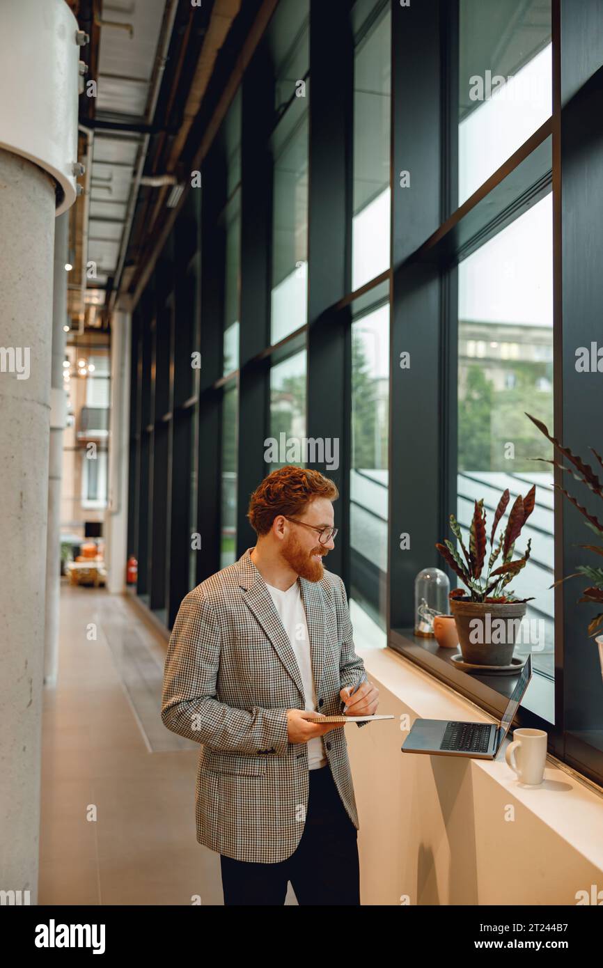 Handsome businessman working on project while using laptop and making notes while standing in office Stock Photo