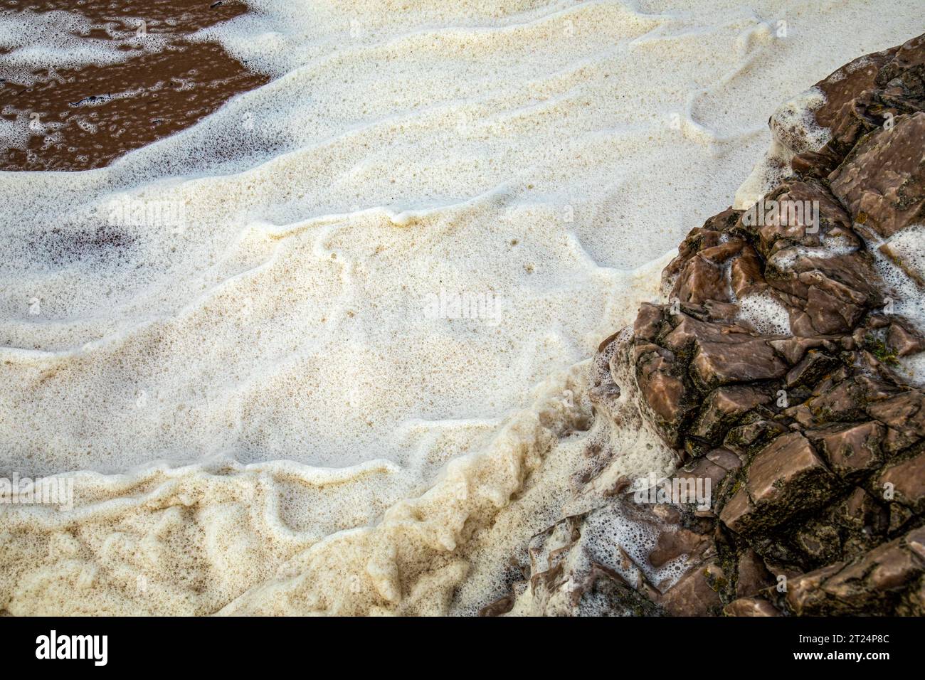 Sea foam whipped up in waves during a storm over rocks and sand on the ...