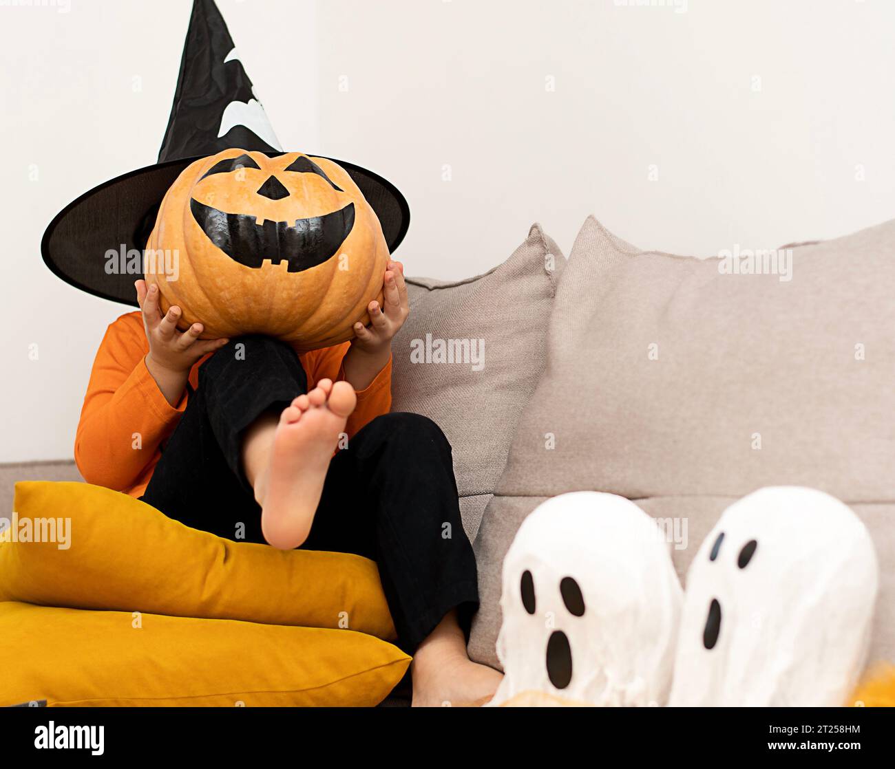 Halloween. Concept. A boy in an orange sweater and a haunted wizard's hat, with a pumpkin instead of a face, sits cheerfully in a home interior. Stock Photo