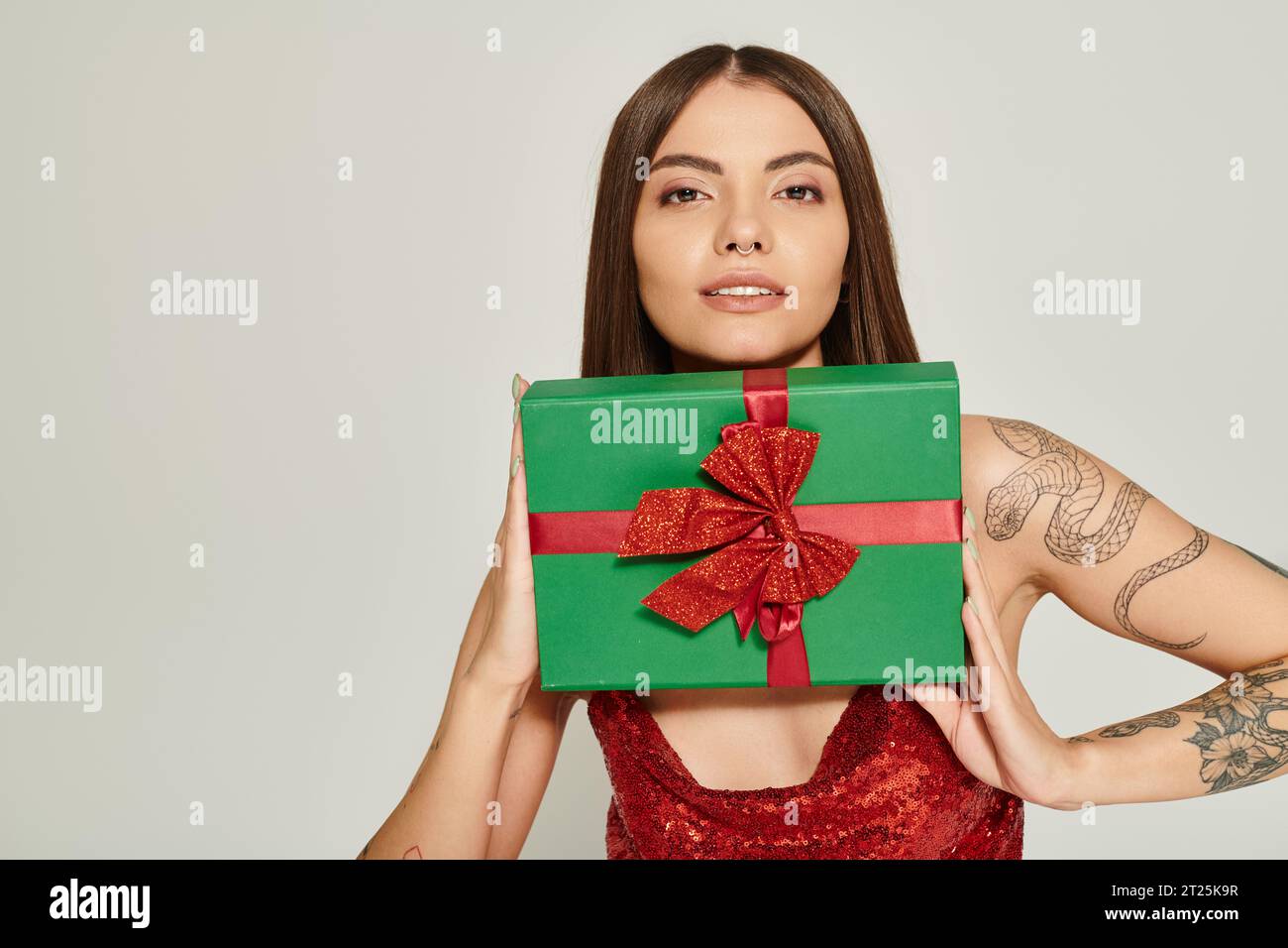 jolly woman with pierced nose holding present under her chin on ecru backdrop, holiday gifts concept Stock Photo
