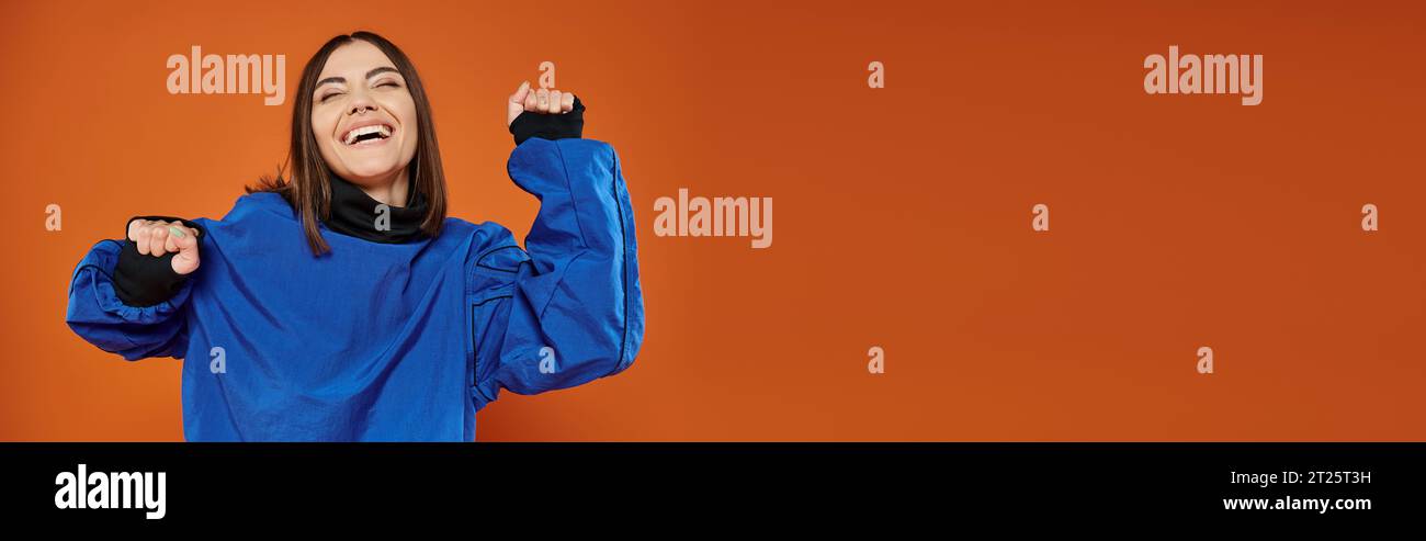 banner of excited woman with pierced nose gesturing and smiling on orange backdrop, blue sweatshirt Stock Photo