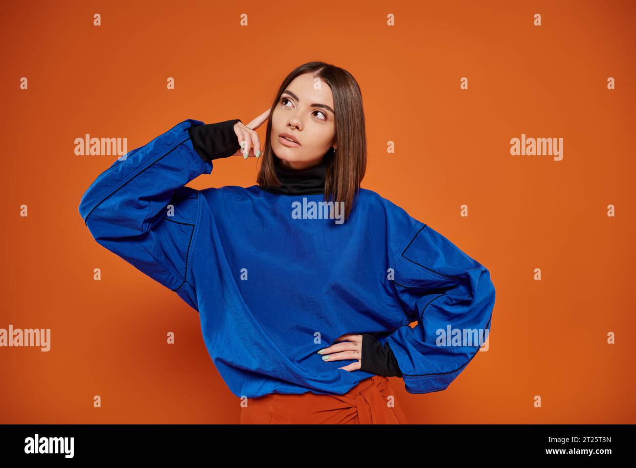 pensive young woman with pierced nose standing with hand on hip on orange backdrop, look away Stock Photo