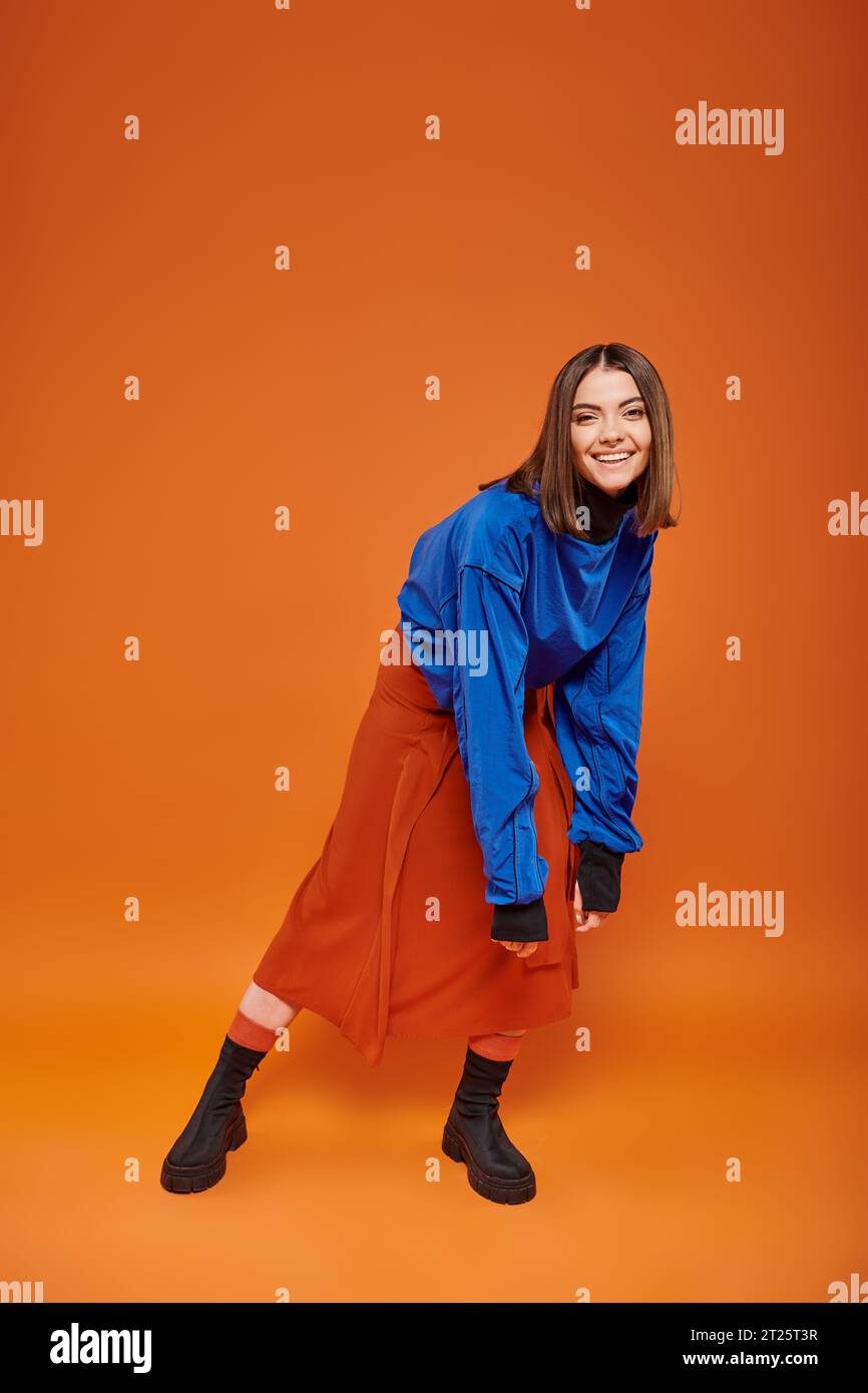 full length of happy young woman with pierced nose standing in autumn attire on orange backdrop Stock Photo