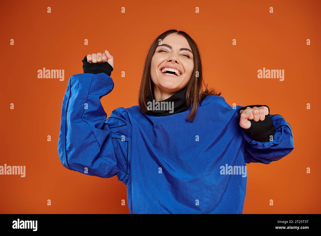 excited young woman with pierced nose gesturing and smiling on orange backdrop, blue sweatshirt Stock Photo