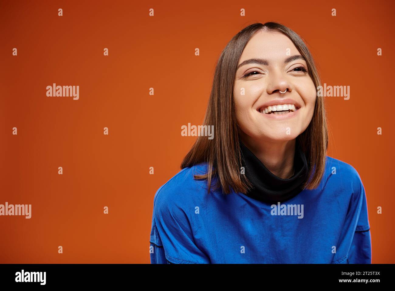 happy young woman with pierced nose looking away and smiling on orange backdrop, blue sweatshirt Stock Photo