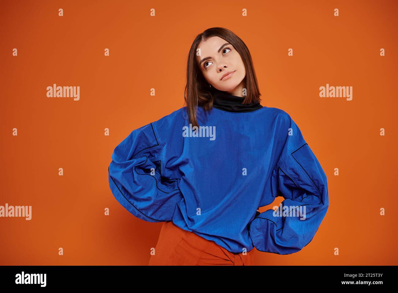 displeased young woman with pierced nose standing with hands on hips on orange backdrop, look away Stock Photo