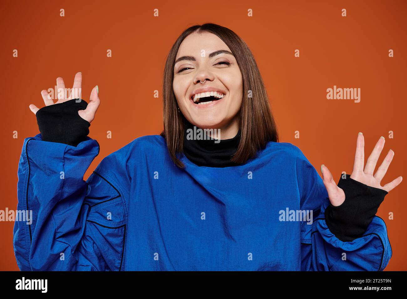 excited young woman with pierced nose looking at camera and smiling on orange backdrop, blue jacket Stock Photo