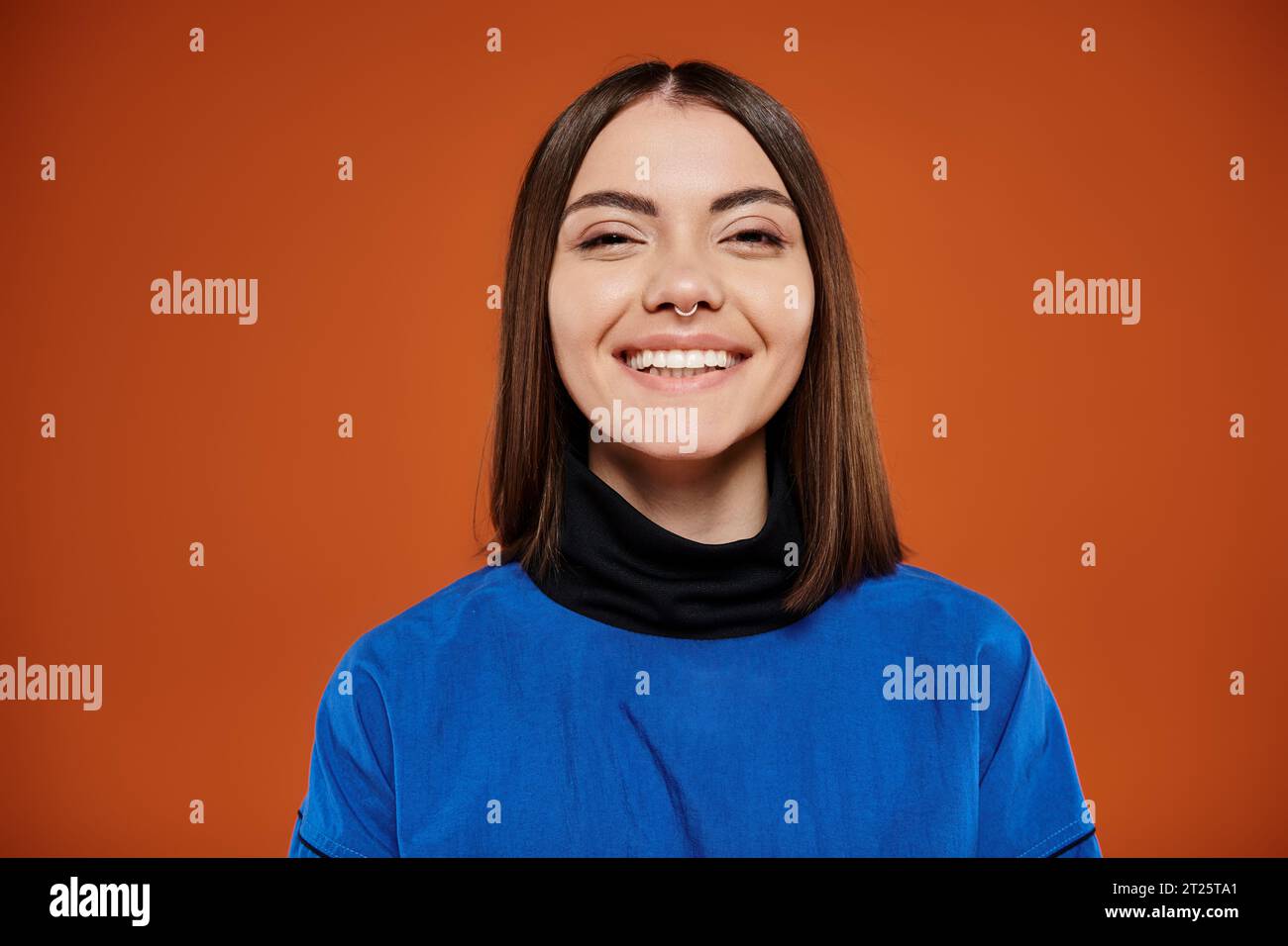 happy young woman with pierced nose looking at camera and smiling on orange backdrop, blue jacket Stock Photo