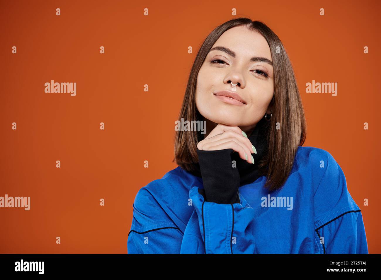pensive woman with pierced nose looking at camera while thinking on orange backdrop, blue jacket Stock Photo
