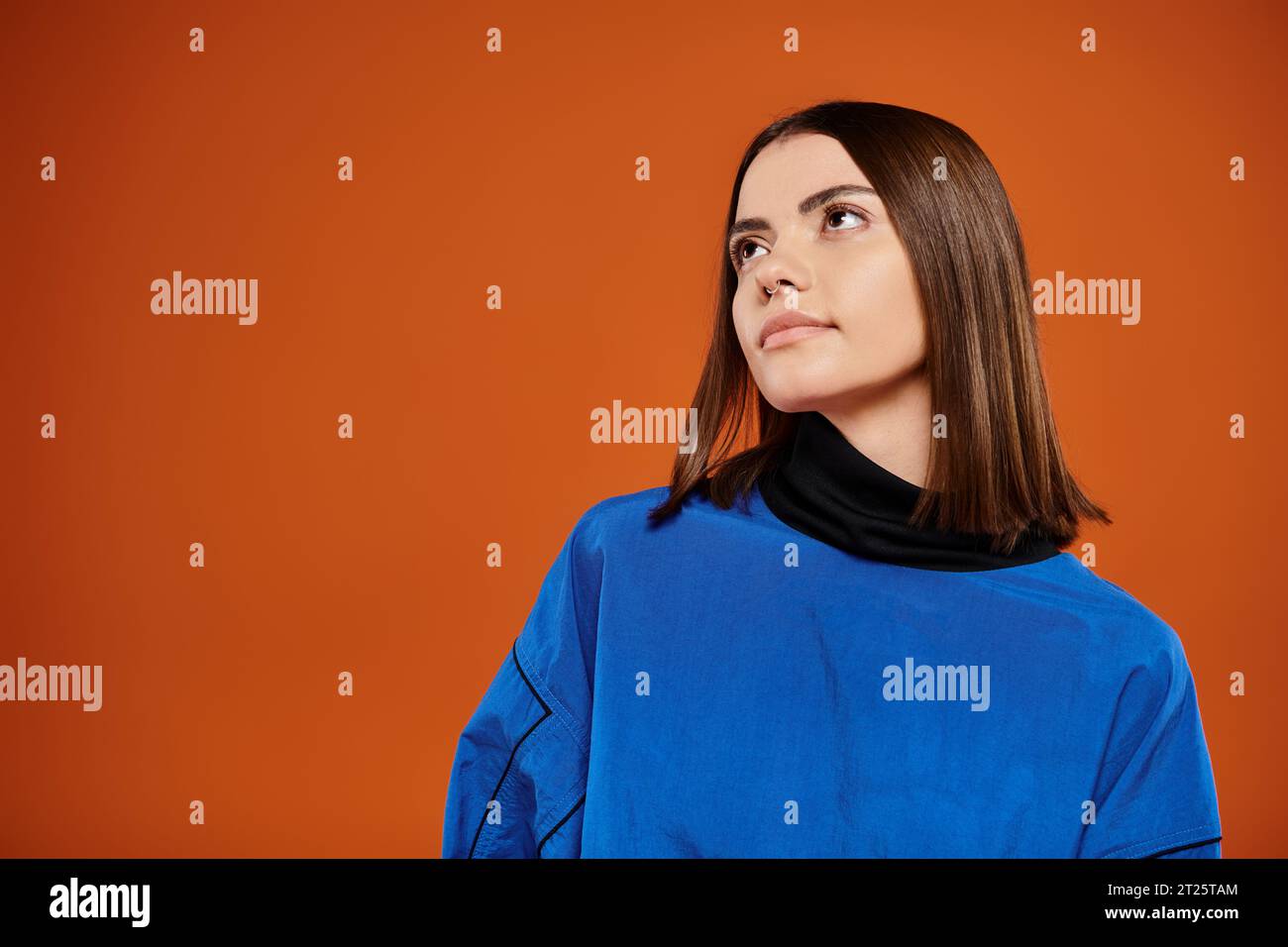 pensive woman with pierced nose looking away while standing in blue jacket on orange background Stock Photo