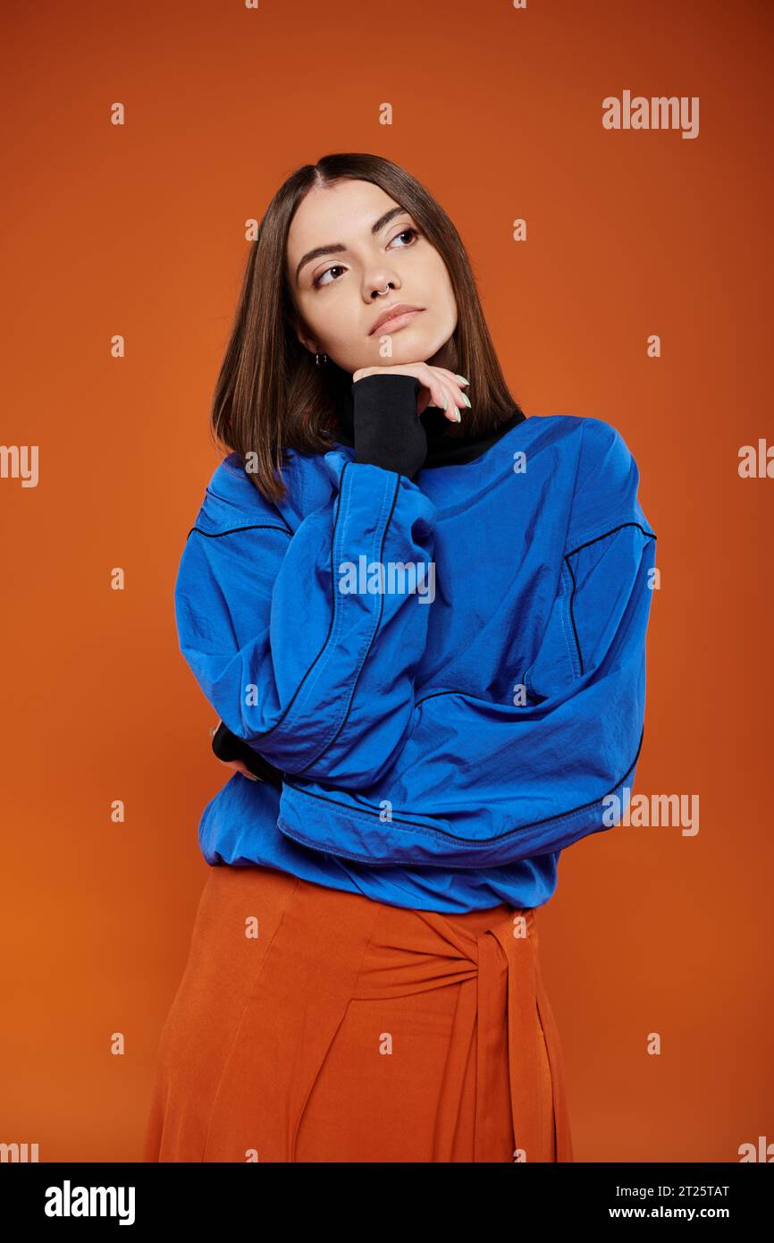 pensive young woman with pierced nose looking away while thinking on orange backdrop, blue jacket Stock Photo