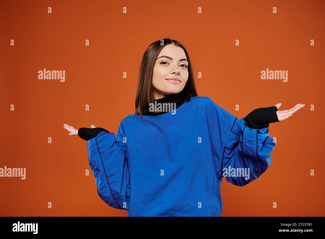 confused woman with pierced nose looking at camera while standing on orange backdrop, blue jacket Stock Photo