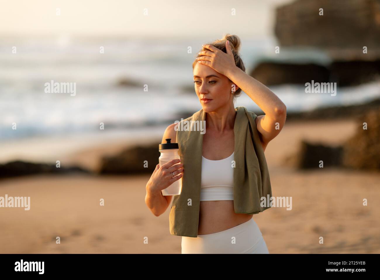 Tired european woman holding bottle, touching head at beach after training Stock Photo