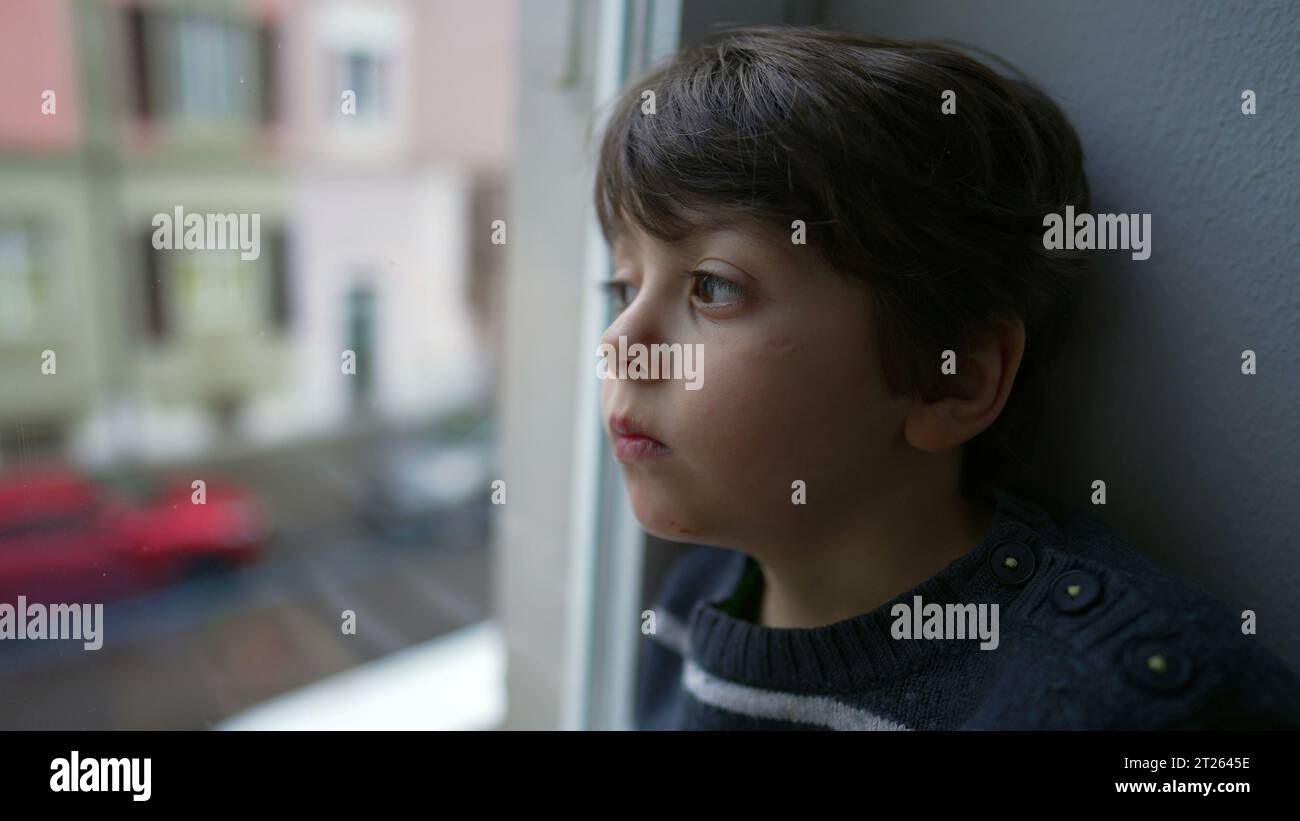 Sad and Bored Little Boy Sitting by Window, Staring in Melancholy, Wanting to Go Out but Stuck at Home Stock Photo