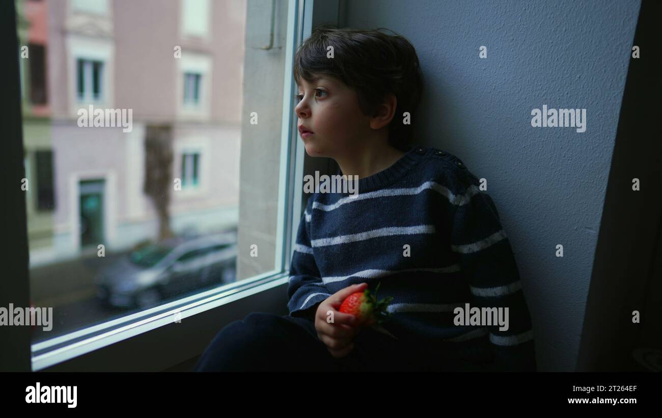 Bored sad emotion of little boy sitting by window staring at view in melancholy, child wanting to go out stuck at home Stock Photo