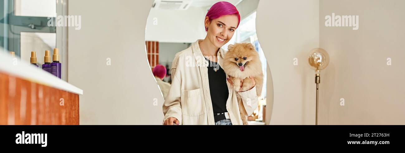 stylish woman with pomeranian spitz smiling at camera near reception desk in dog hotel, banner Stock Photo