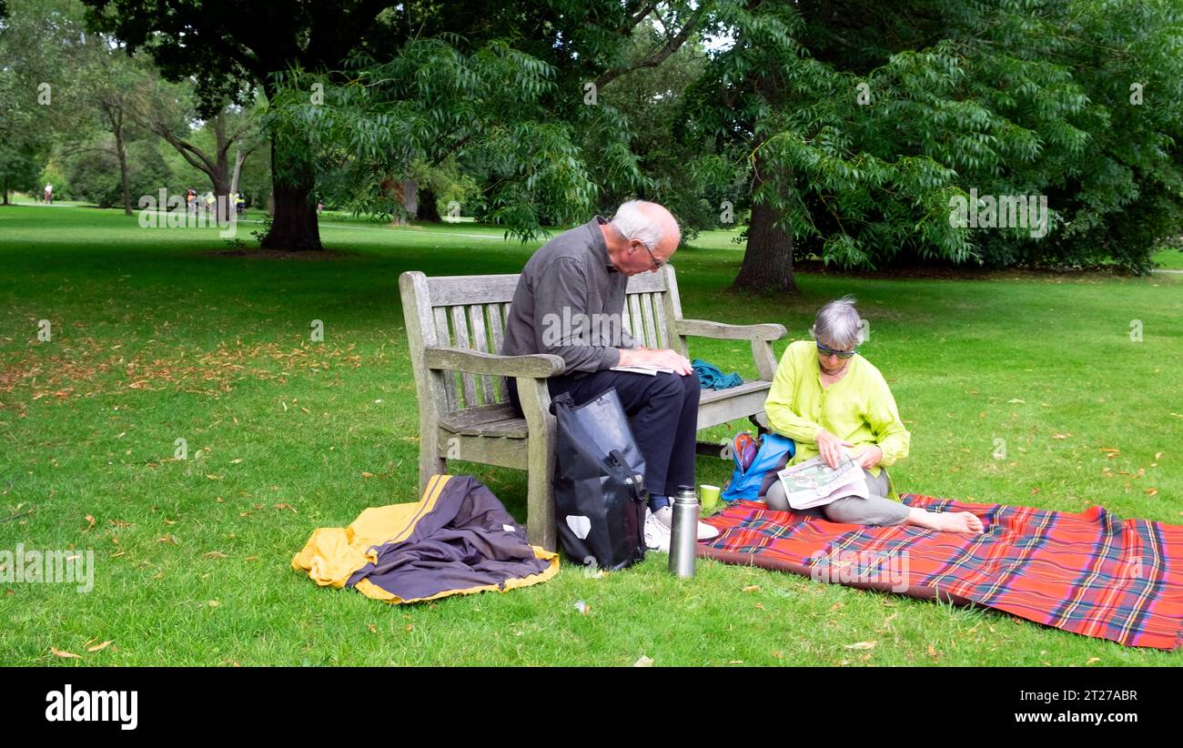 Older couple man woman sitting on wooden garden bench and picnic blanket reading newspaper and drinking coffee tea thermos in summer Kew Gardens UK Stock Photo