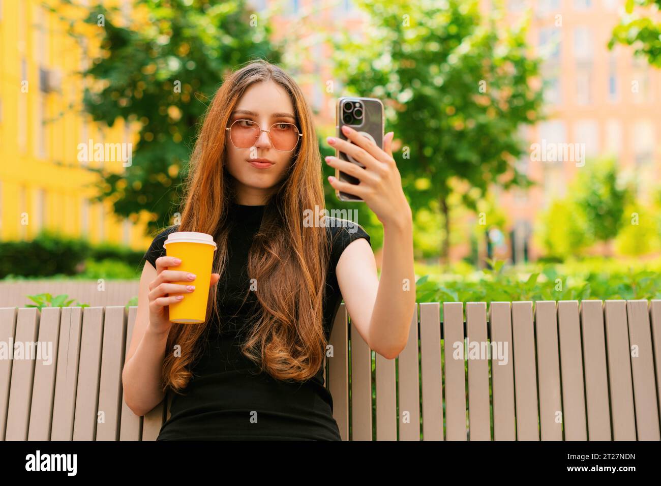 Smiling funny young girl with cool sunglasses using Social media with a Smart Phone sitting on a bench in town a holding disposable paper coffee cup Stock Photo