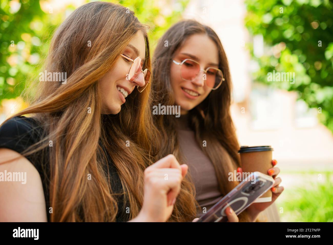 Amazed Twins sisters with sunglasses using Social media with a Smart Phone sitting on a bench in town a holding disposable paper coffee cup outdoors Stock Photo
