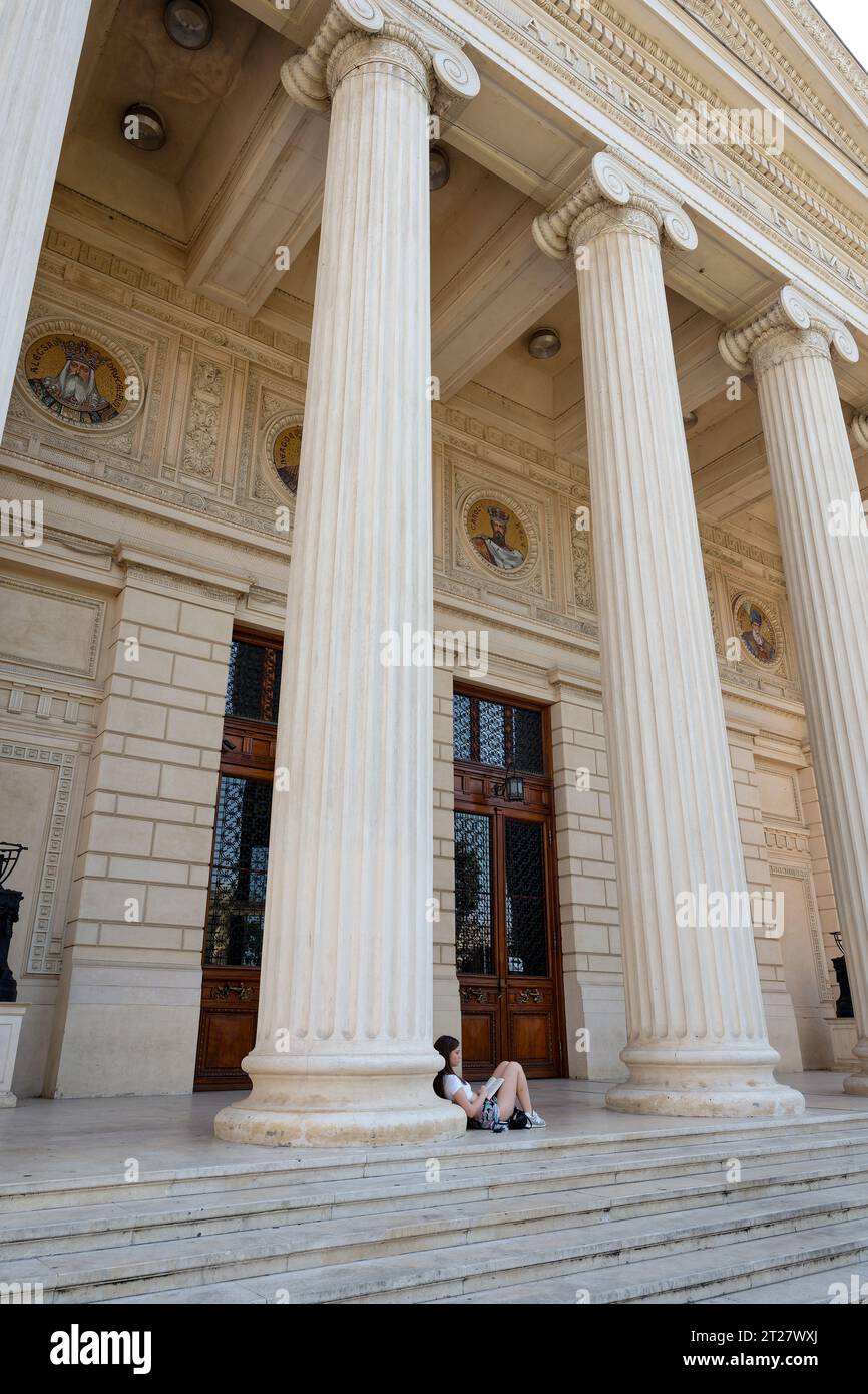Teenager sits serenely, immersed in a book beneath a pillar of the romanian athenaeum in Romania Stock Photo
