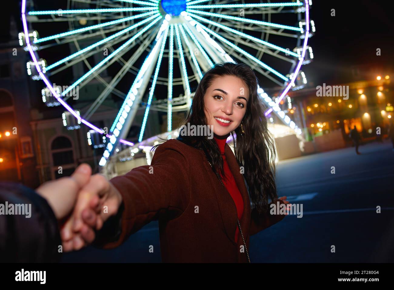 A young beautiful woman in a red coat takes her boyfriend's hand while walking in front of a Ferris wheel at an amusement park. Stock Photo