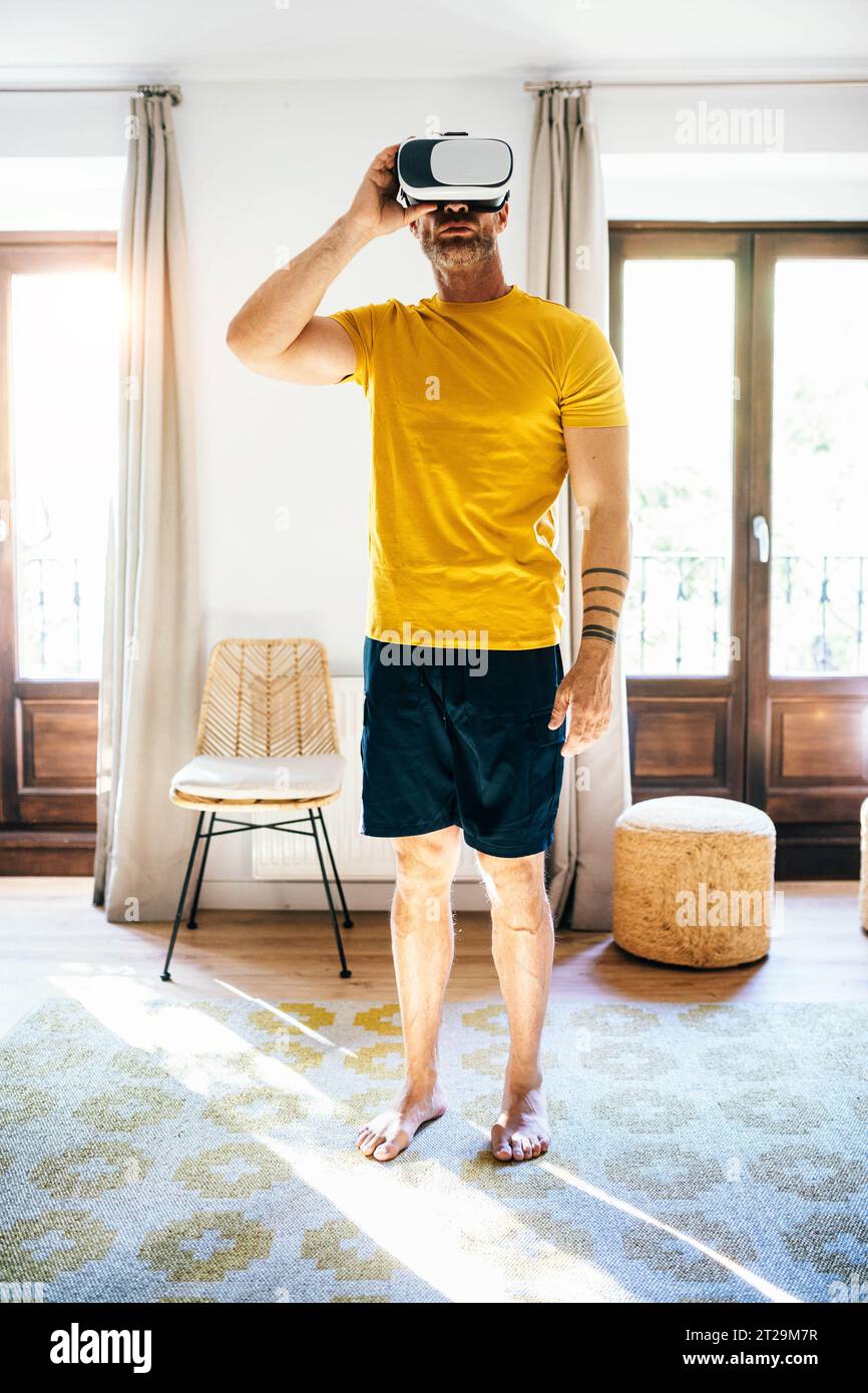 Full body of barefoot man in activewear looking through VR glasses while standing in cozy room in sunlight Stock Photo