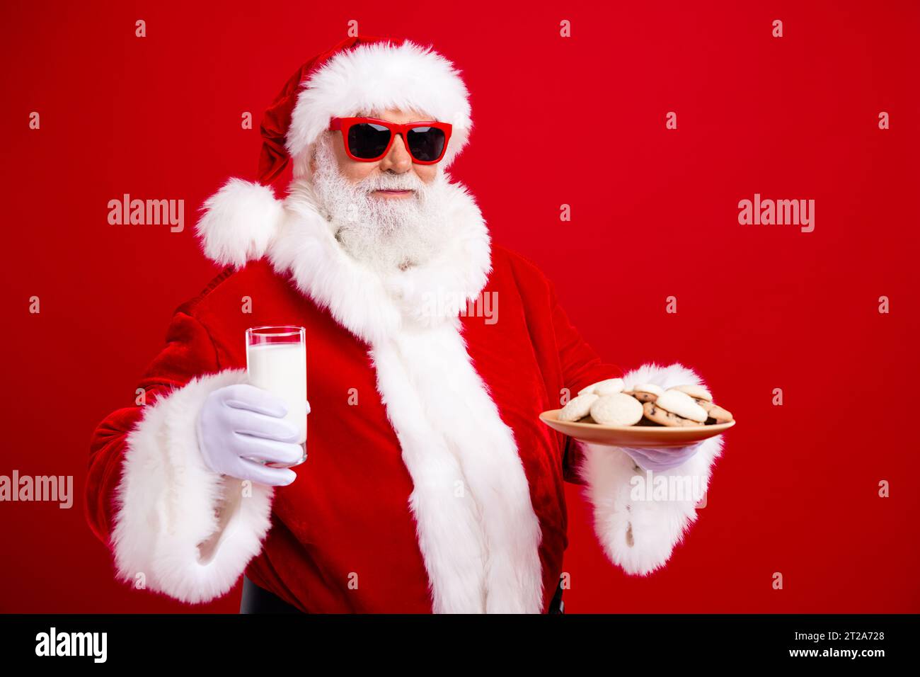 Photo of positive cheerful grandfather wear stylish santa costume hold glass of milk cookies in hands isolated on red color background Stock Photo