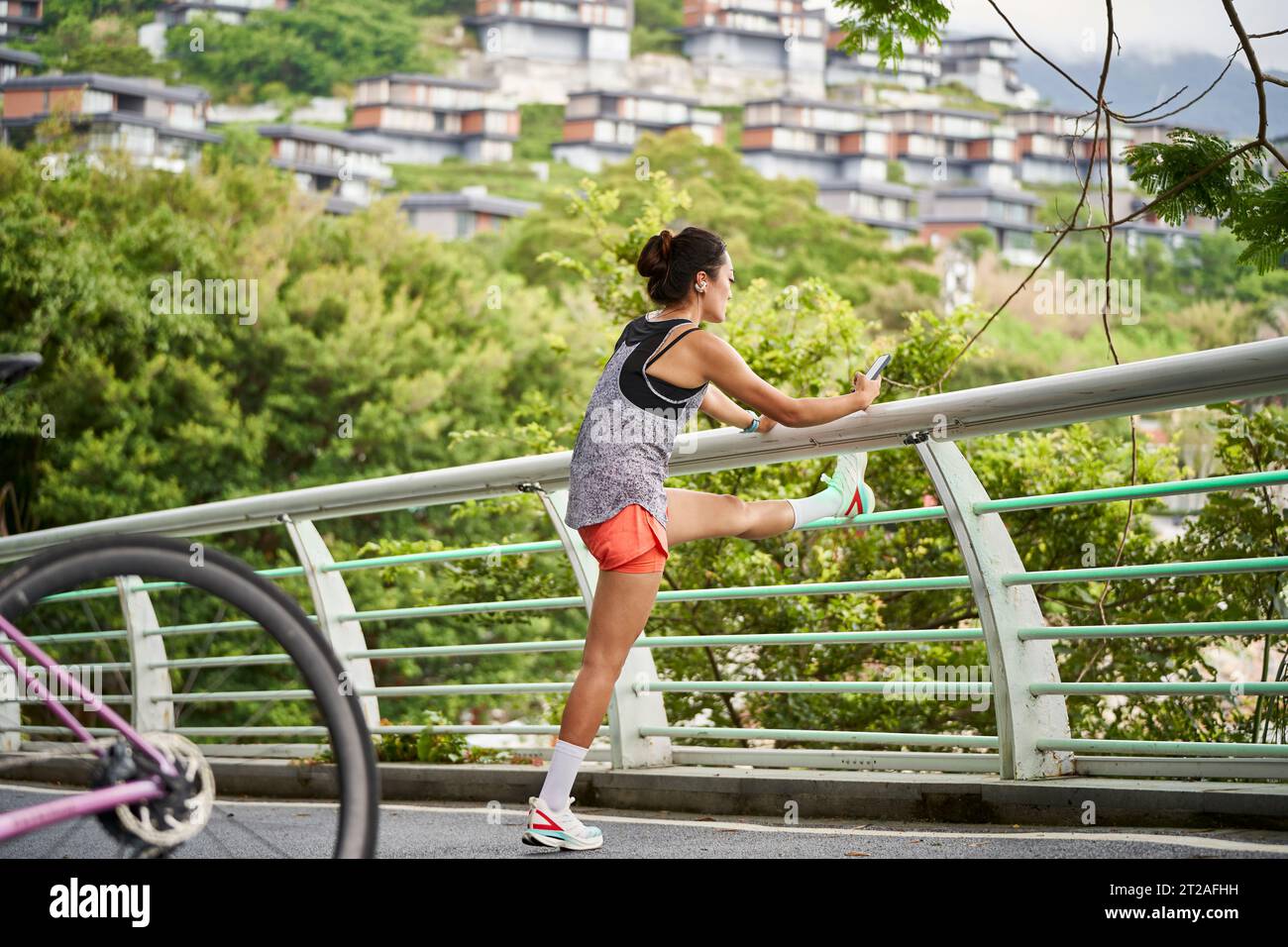 young asian woman looking at cellphone while warming up by pressing legs outdoors Stock Photo