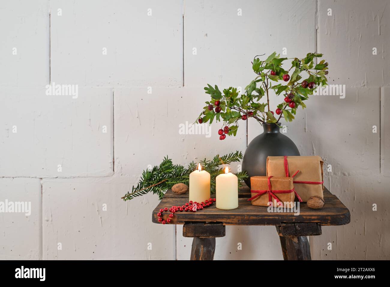 Natural Advent and Christmas decoration, gifts, candles and branches with red fruits in a vase on a rustic wooden stool in front of a rough white wall Stock Photo