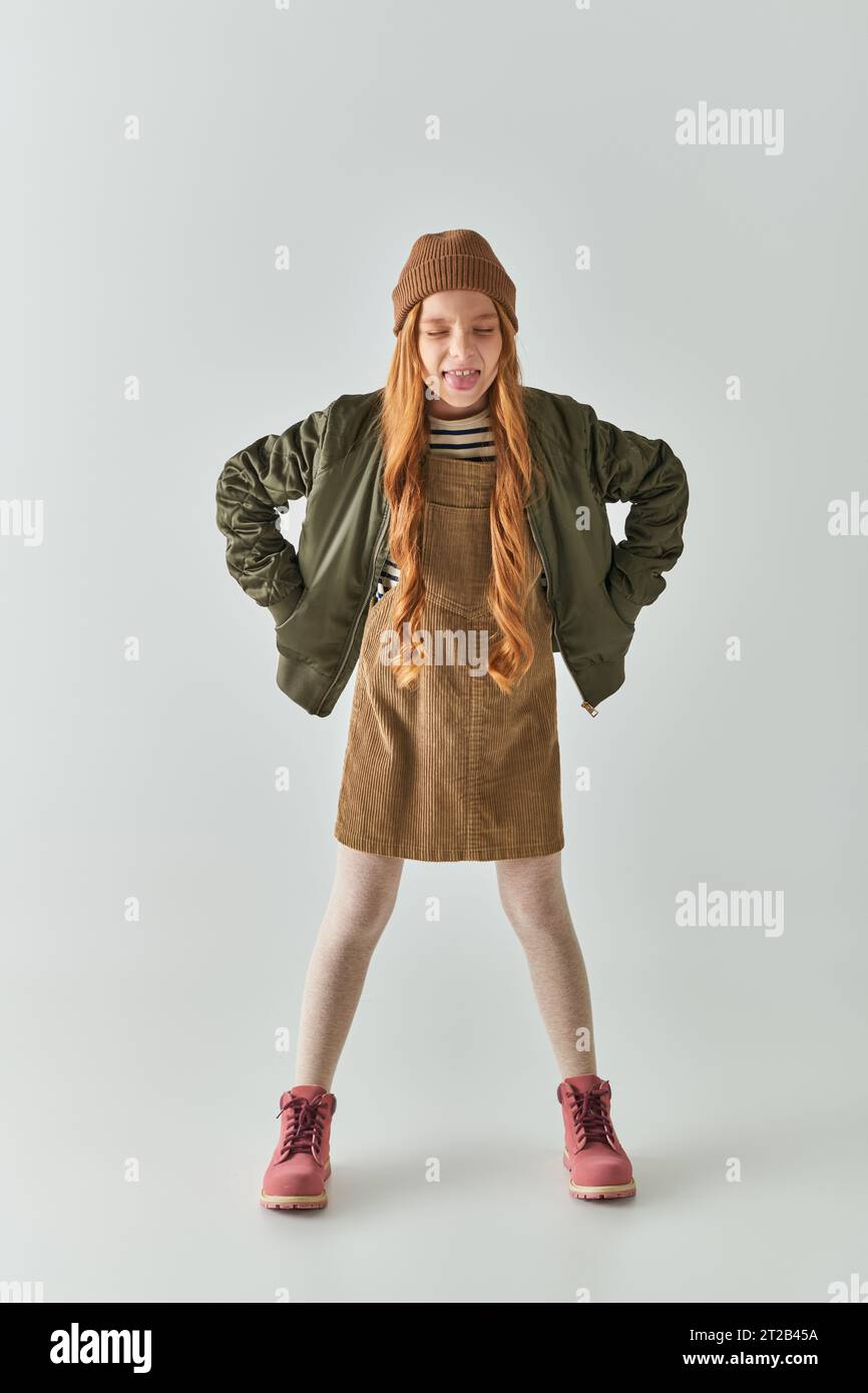 girl with long hair and winter hat on head sticking tongue and standing in dress and jacket on grey Stock Photo