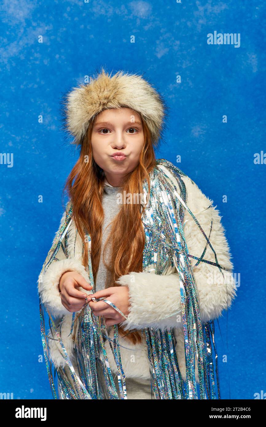 girl in faux fur jacket with tinsel standing under falling snow on blue