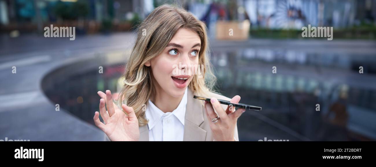 Chatty businesswoman sitting in city centre and recording voice message, talking into microphone on smartphone Stock Photo