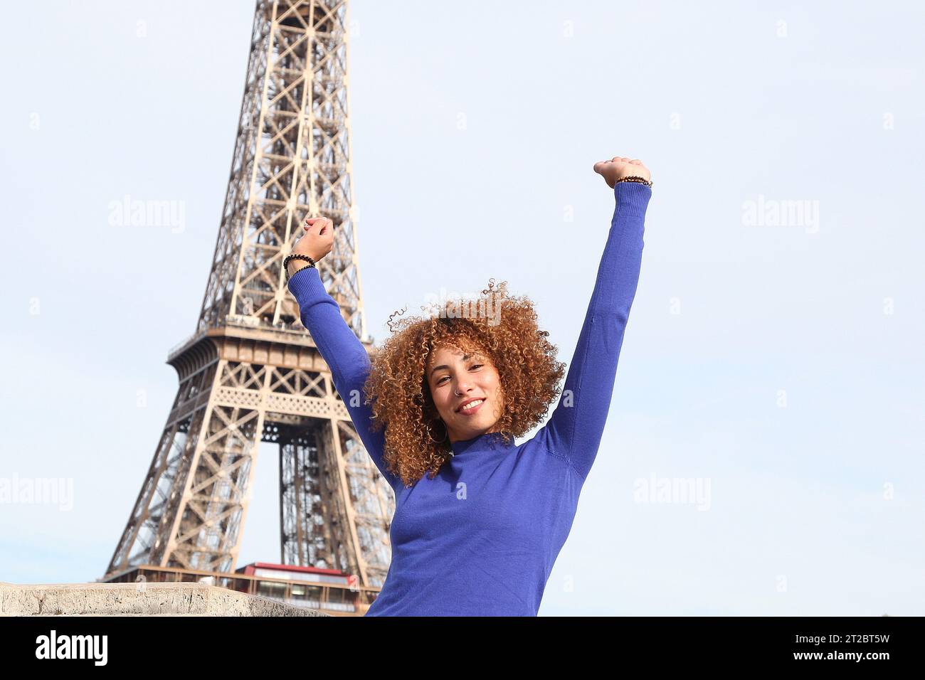HAPPY CURLY HAIR YOUNG WOMAN IN BLUE SWEATER HAVING A GOOD TIME AT THE EIFFEL TOWER Stock Photo