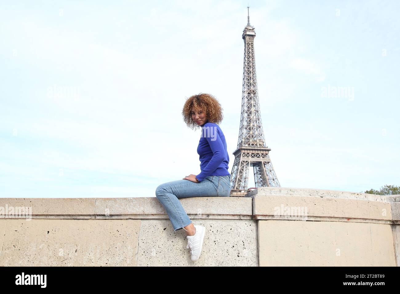 HAPPY CURLY HAIR YOUNG WOMAN IN BLUE SWEATER HAVING A GOOD TIME AT THE EIFFEL TOWER Stock Photo