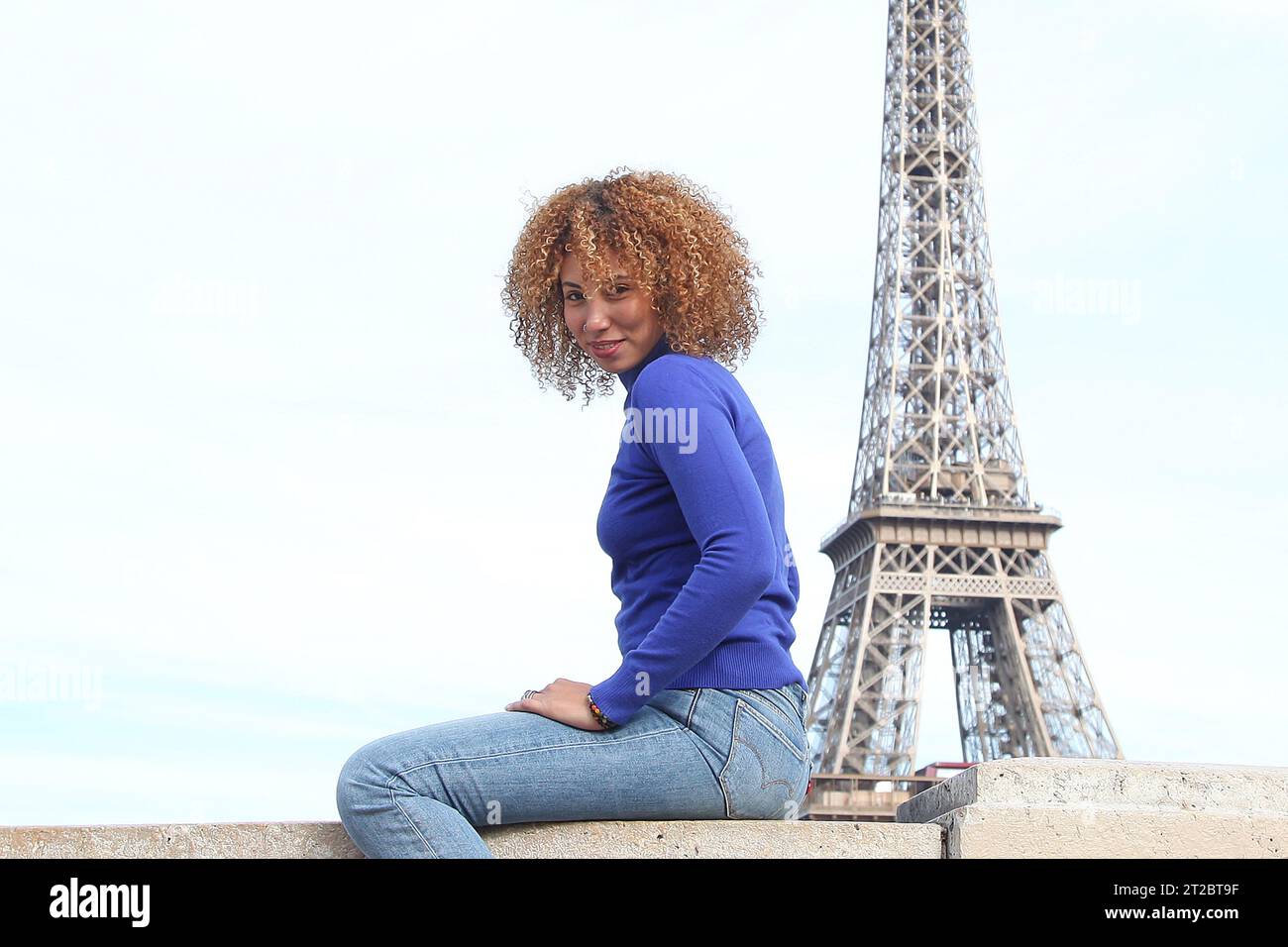 HAPPY CURLY HAIR YOUNG WOMAN IN BLUE SWEATER HAVING A GOOD TIME AT THE EIFFEL TOWER Stock Photo