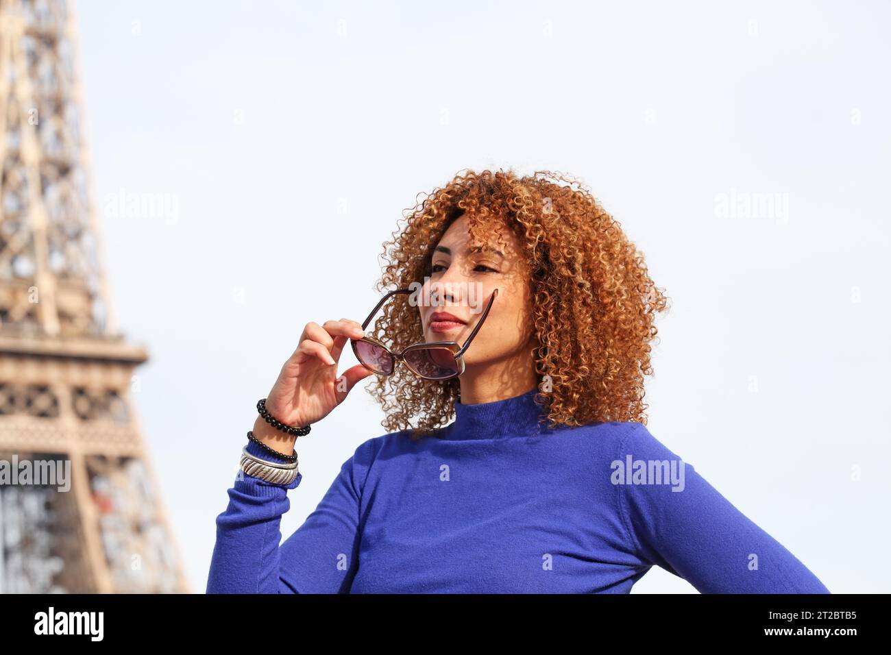 HAPPY CURLY HAIR YOUNG WOMAN IN BLUE SWEATER HOLDING HER SUNGLASSES AT THE EIFFEL TOWER Stock Photo