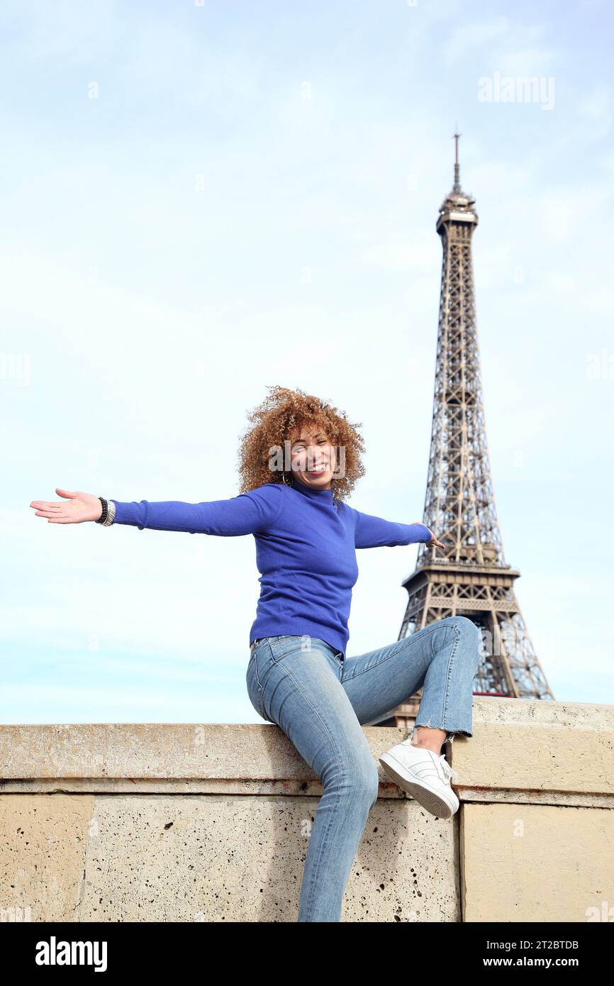 HAPPY CURLY HAIR YOUNG WOMAN IN BLUE SWEATER HAVING A GOOD TIME AT THE EIFFEL TOWER Stock Photo