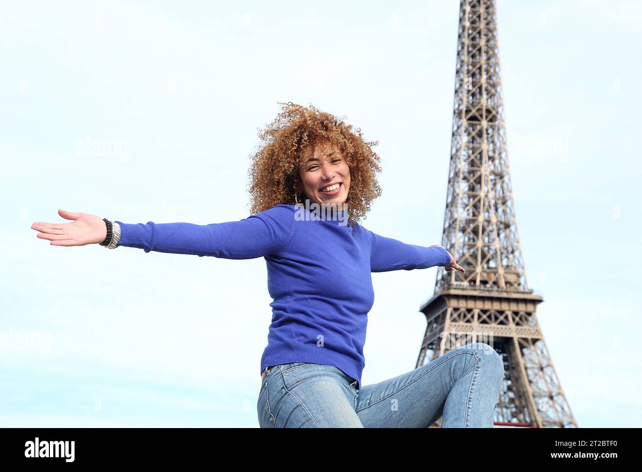 HAPPY CURLY HAIR YOUNG WOMAN IN BLUE SWEATER HAVING A GOOD TIME AT THE EIFFEL TOWER Stock Photo
