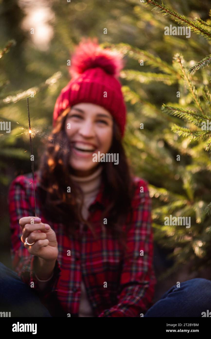 Bright sparkler in hands near defocused background of young woman in red checkered shirt among green pine trees. Curly-haired girl laughs, rejoices at Stock Photo