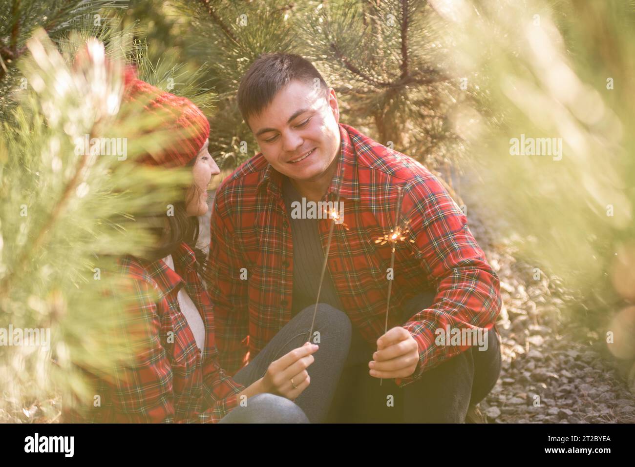 Through green needles of pine trees, young happy couple in red checkered shirts hold festive sparklers in forest. Man and woman in hat laugh at Christ Stock Photo
