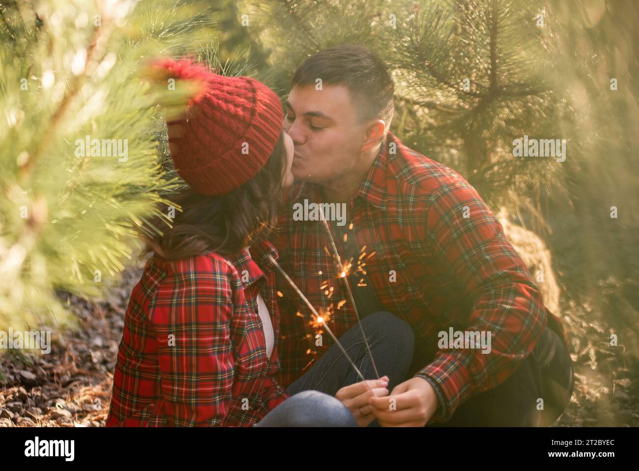 Through green needles of pine trees, young happy couple in red checkered shirts hold festive sparklers in forest. Man and woman in hat kissing at Chri Stock Photo