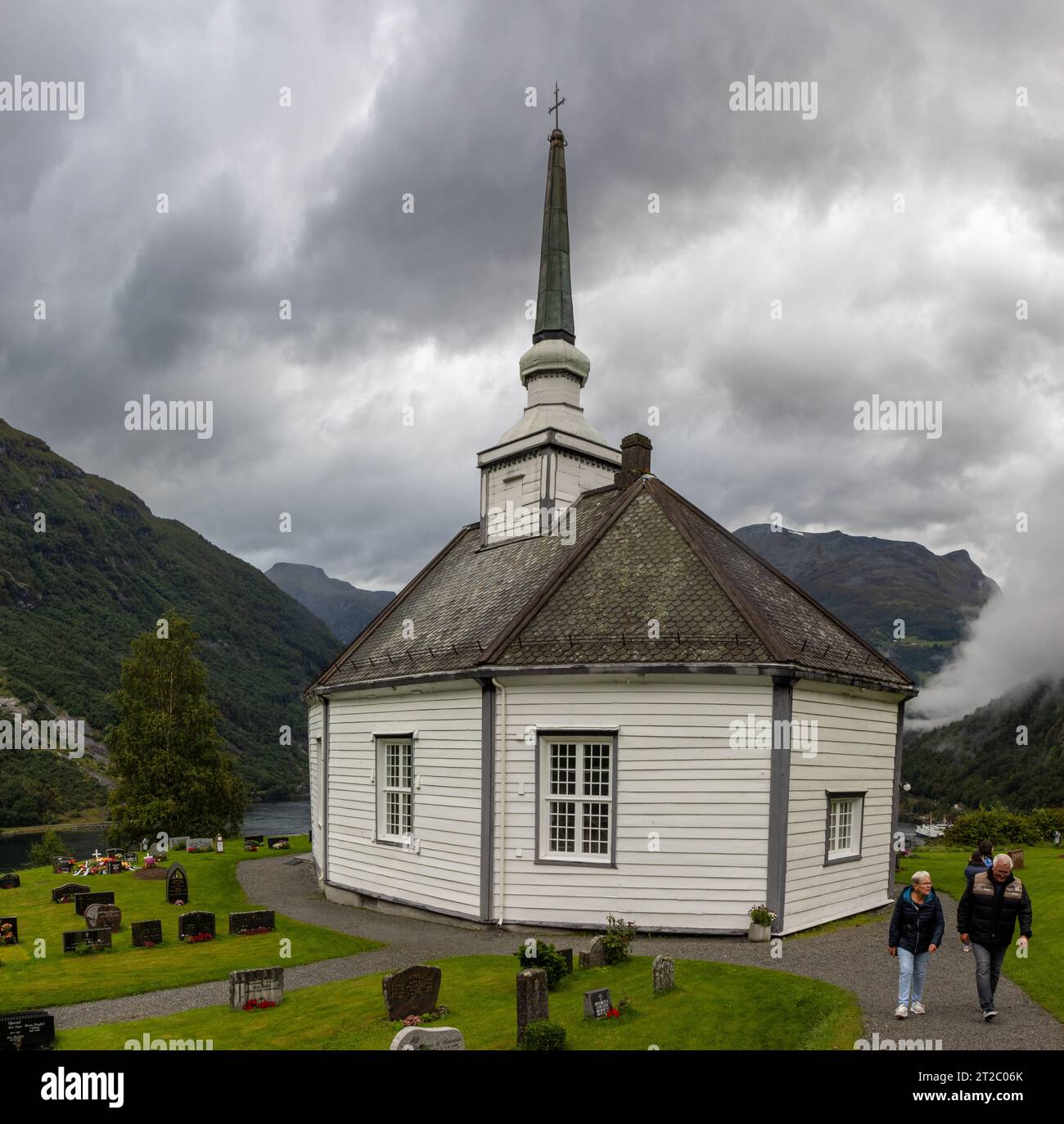 Pareja de turistas mayores visitando la iglesia octogonal de Geiranger. Está hecha de madera blanca y tejado de pizarra negra, Noruega Stock Photo