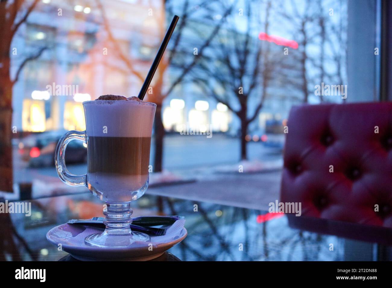 A cup of hot, brown tasty cappuccino with milk on a saucer divided into layers with a straw on the table in a cafe in the evening against the window. Stock Photo