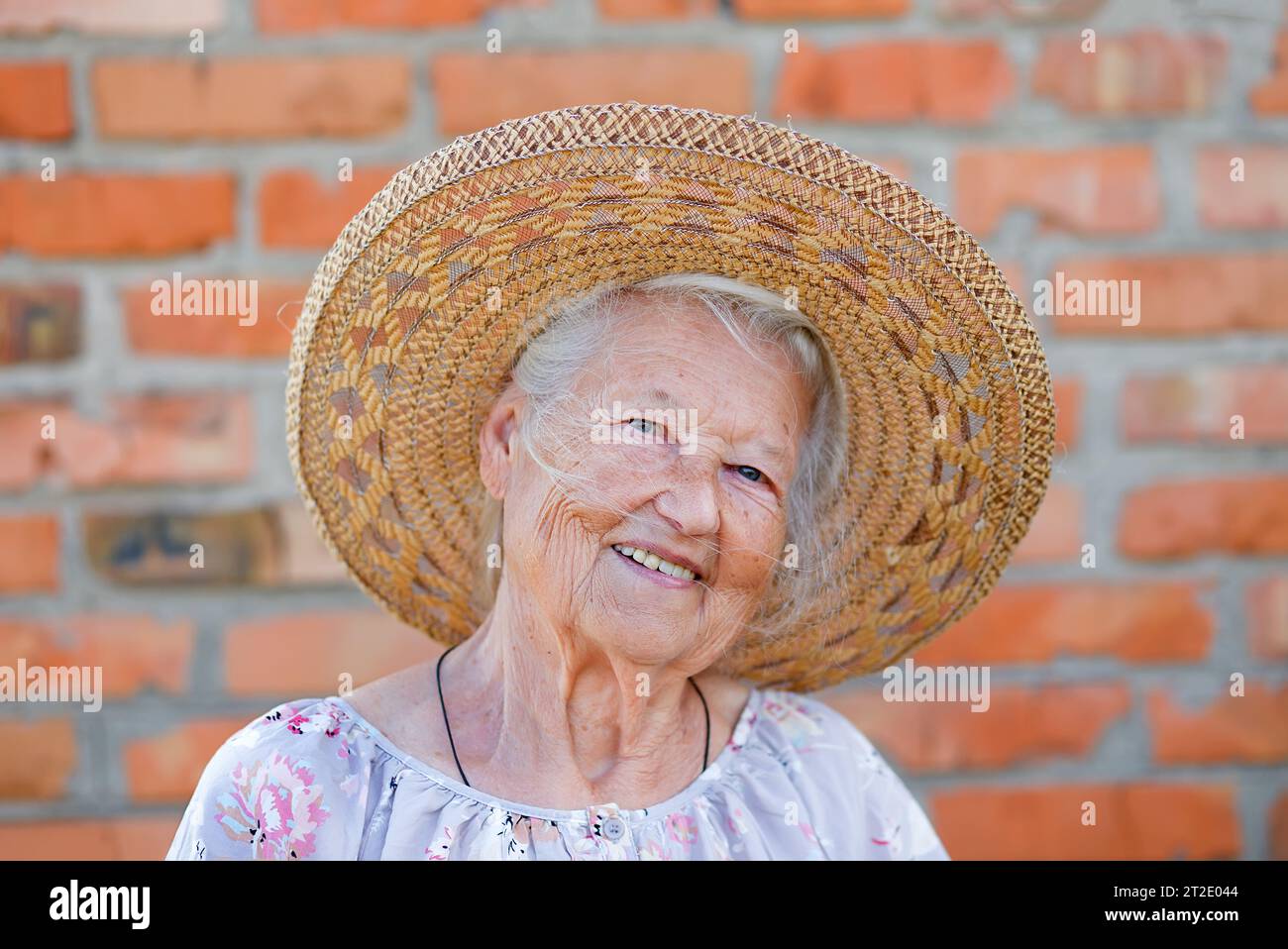 Senior woman portrait, on red brick wall background with white hair.Happy smiling grandma in straw hat Stock Photo