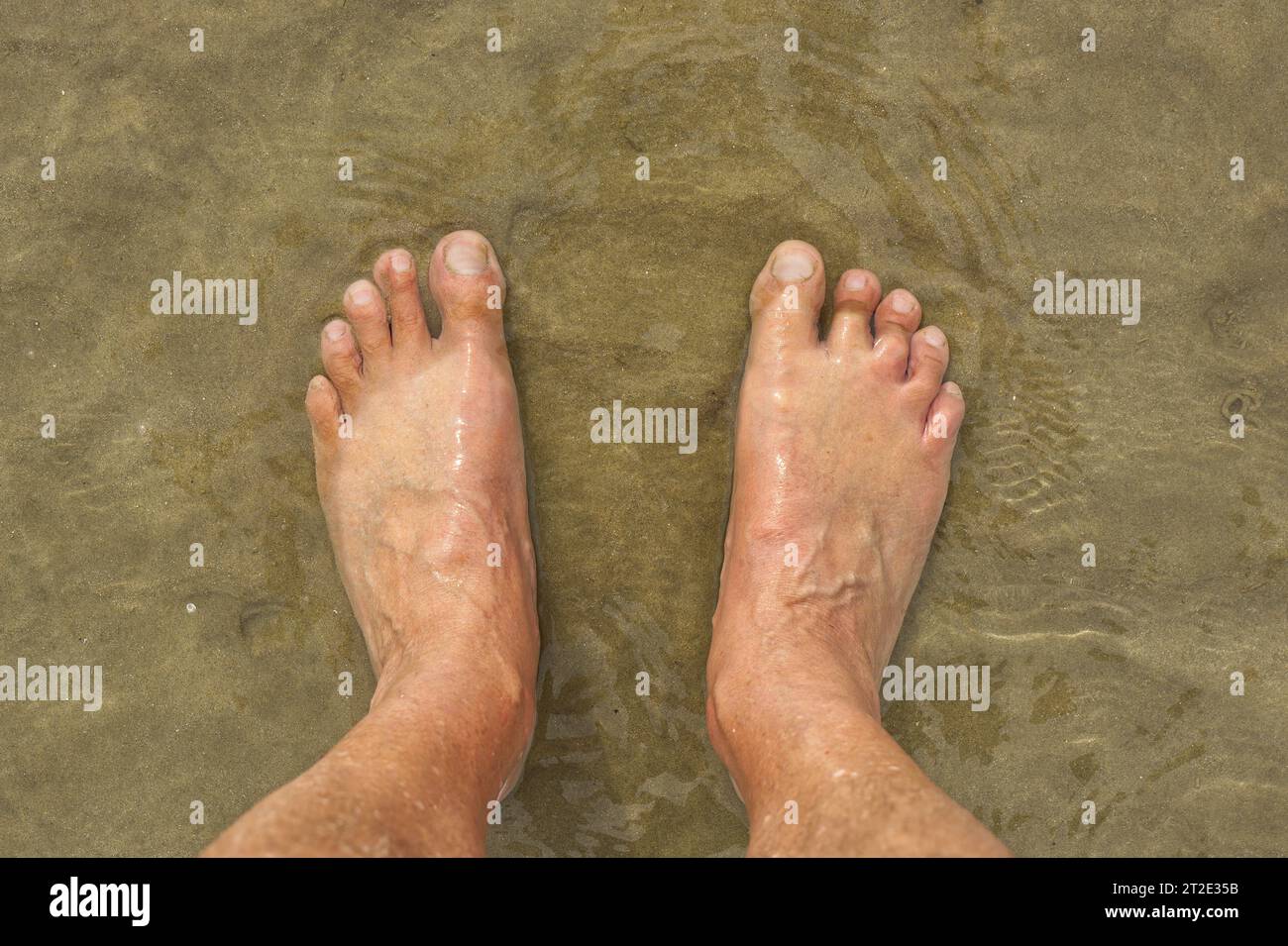 Pair of adult male feet on sandy beach with clear water washing over. Stock Photo