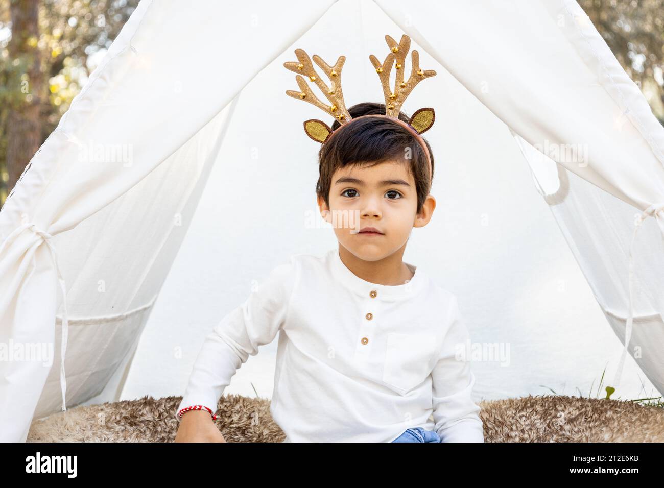 Smiling child boy playing with Christmas decoration in outdoor teepee tent with glitter reindeer antlers. Winter family photoshoot in the forest. Stock Photo