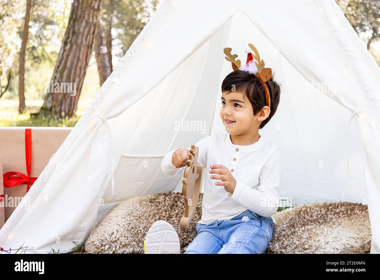 Smiling child boy playing with Christmas decoration in outdoor teepee tent with glitter reindeer antlers. Winter family photoshoot in the forest. Stock Photo